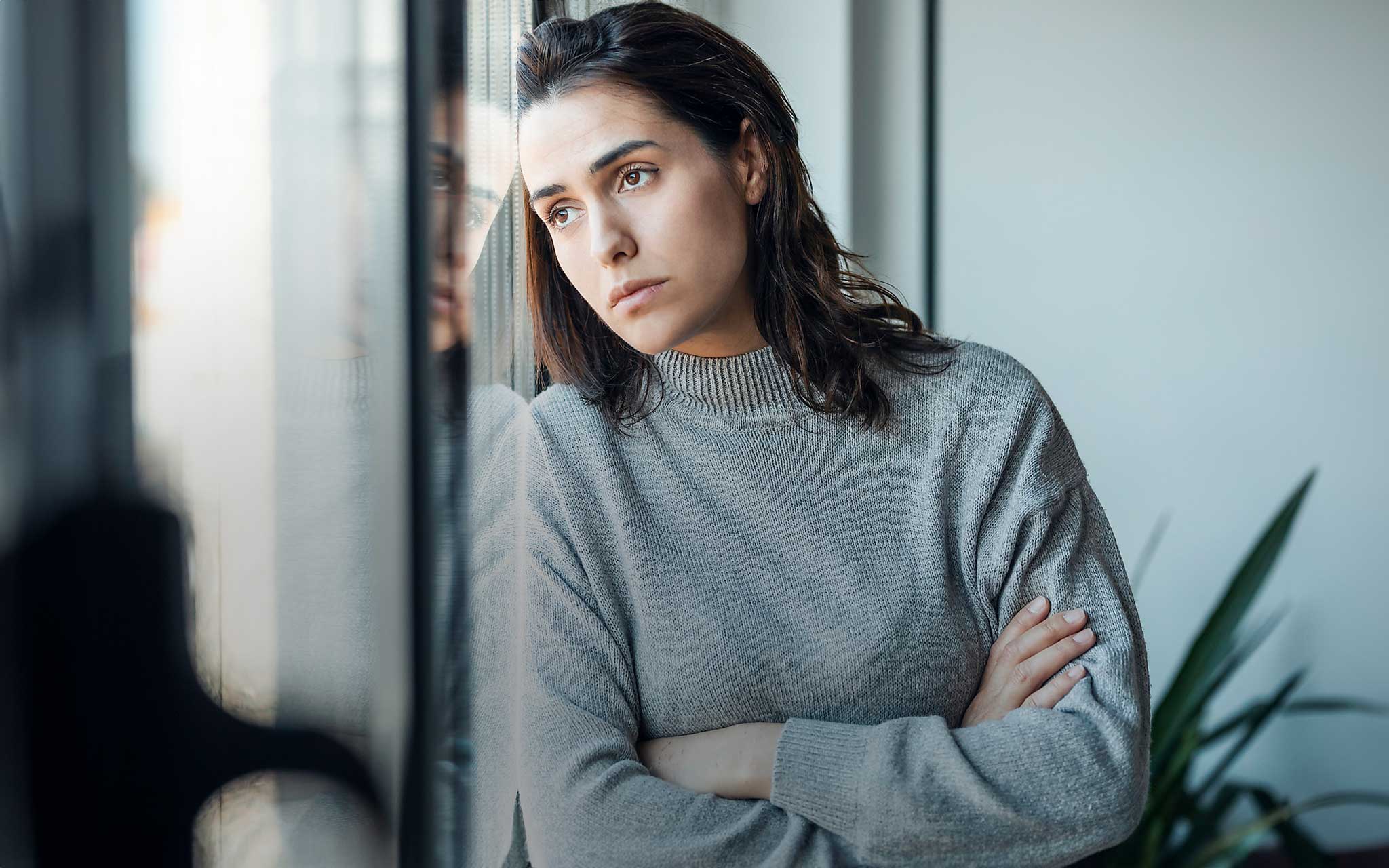Traurige Frau steht mit verschränkten Armen und schaut durch das Fenster zu Hause