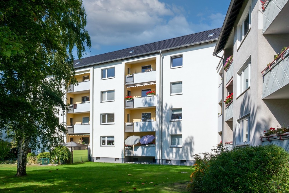 Mehrstöckige Wohngebäude mit Balkonen, grüner Rasenfläche und Bäumen im sonnigen Außenbereich.