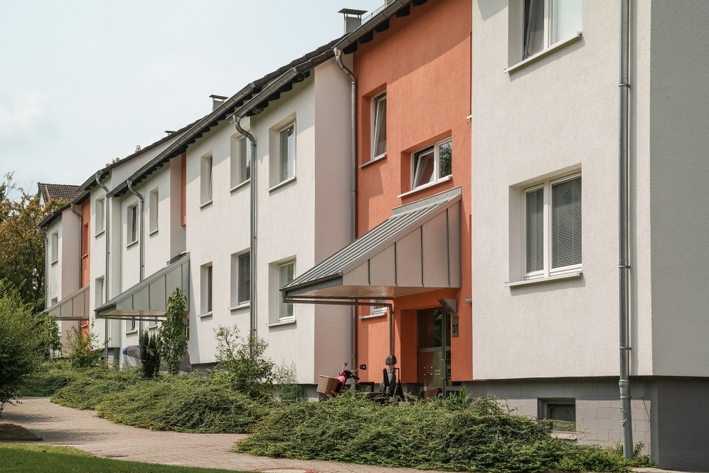 Reihenhäuser mit weißen und orangefarbenen Fassaden und begrünten Vorgärten bei Tag.