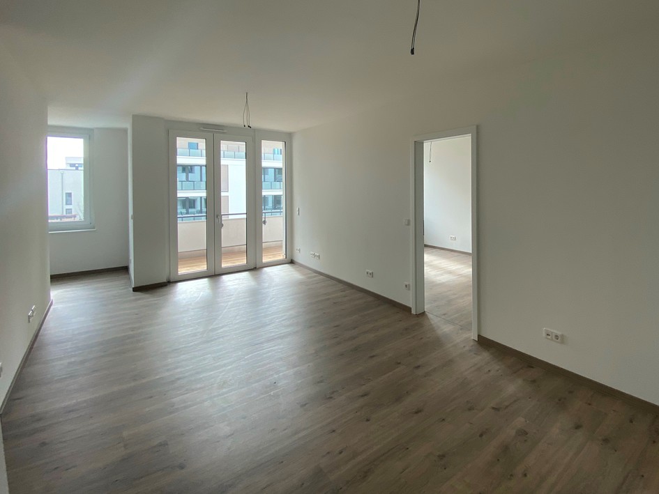 Helle, leere Wohnung mit Holzboden, Balkonzugang durch Glasfenster und angrenzendem Raum.