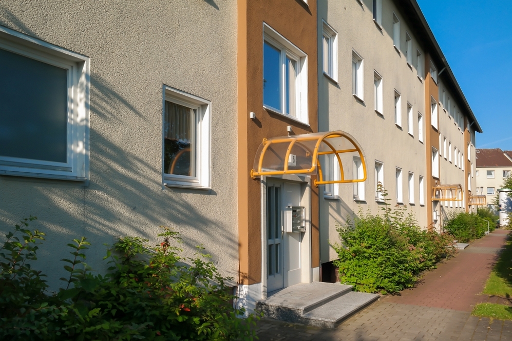 Mehrfamilienhaus mit Eingängen und gelben Vordächern, umgeben von Grün und Gehweg.