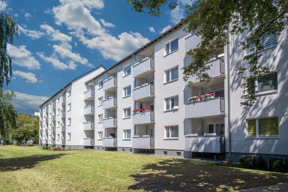 Weiße Mehrfamilienhäuser mit Balkonen und Blumen vor grünem Rasen unter blauem Himmel.