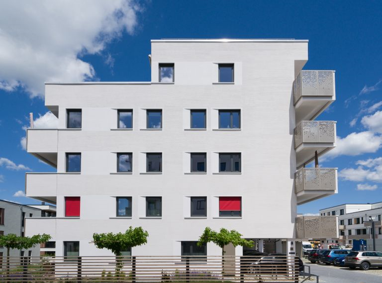 Modernes weißes Mehrfamilienhaus mit Balkonen und roten Fensterläden bei klarem Himmel.