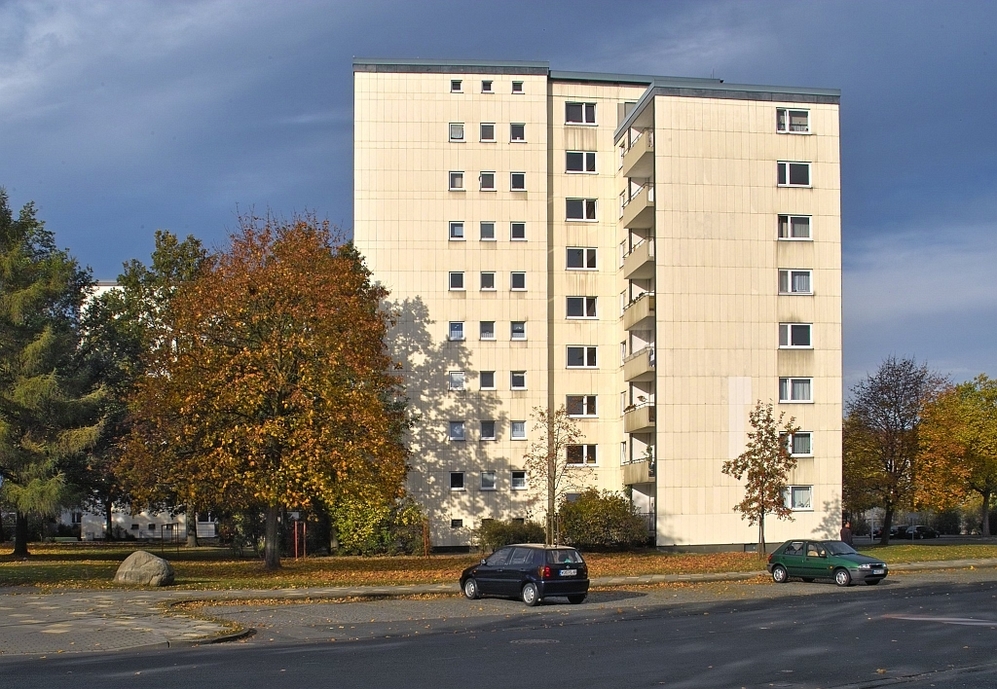 Neun-geschossiges Wohnhaus mit herbstlichen Bäumen und zwei parkenden Autos davor.