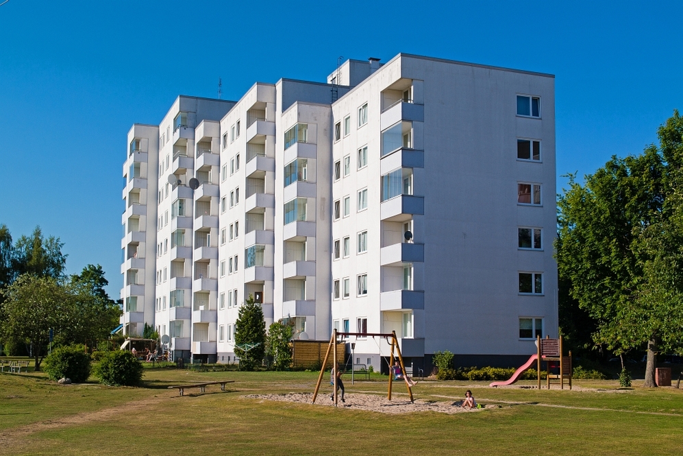 Mehrstöckiges weißes Wohngebäude mit Balkonen und Spielplatz im Vordergrund bei blauem Himmel.