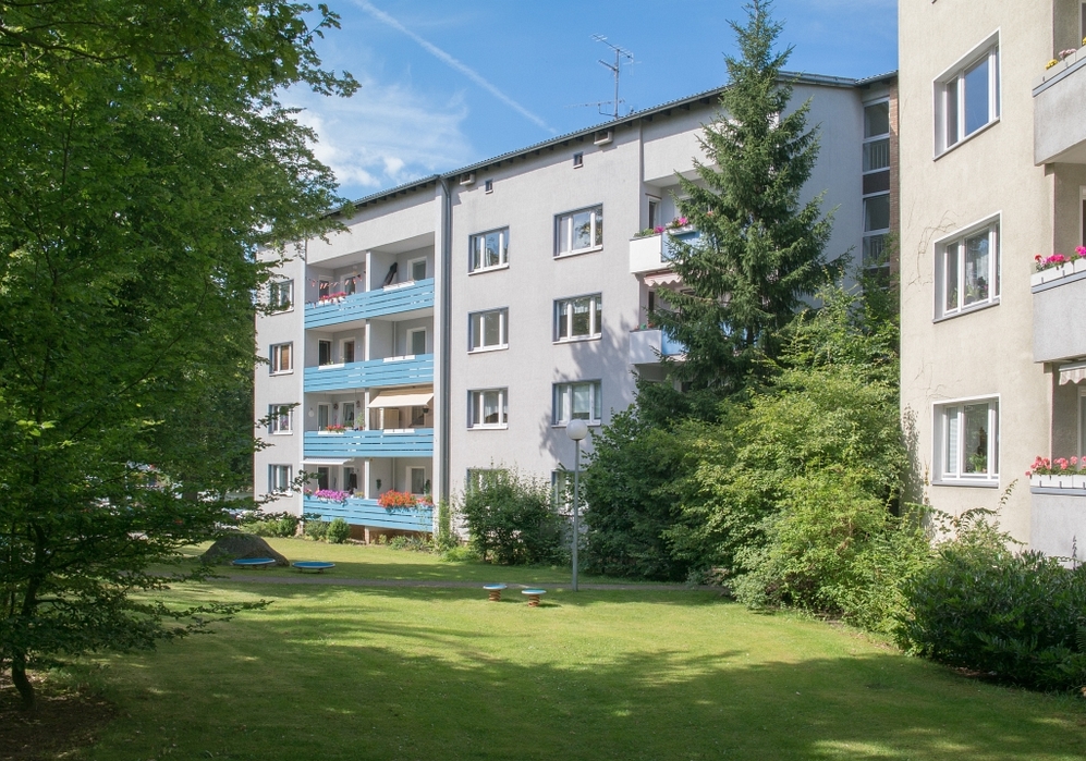Mehrfamilienhaus mit blauen Balkonen und gepflegtem Garten bei Sonnenschein.