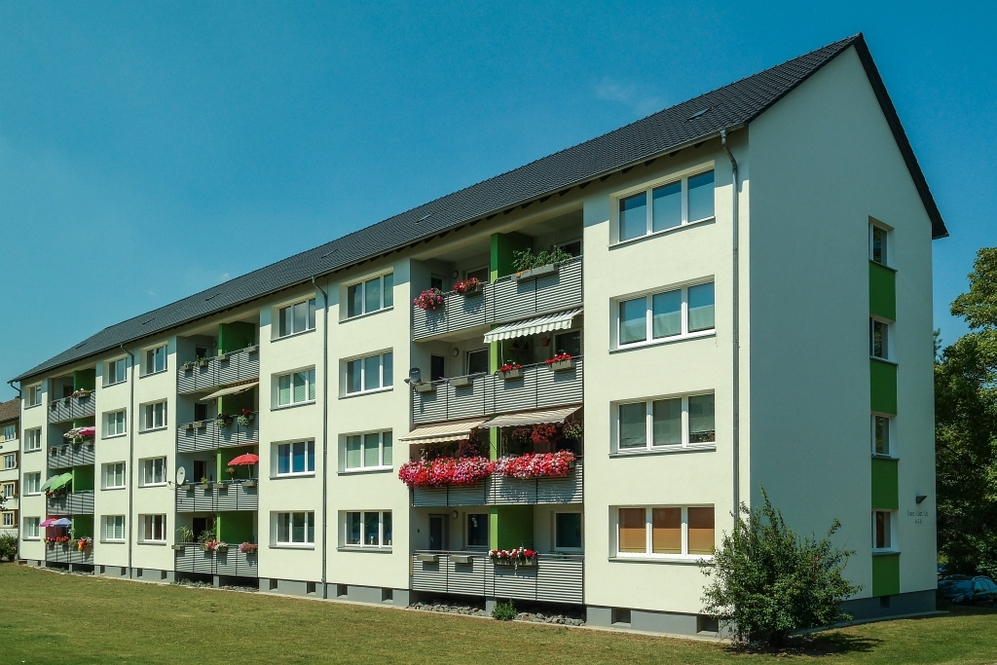 Mehrstöckiges Wohngebäude mit Balkonen und Blumen, grüner Wiese im Vordergrund, blauer Himmel.