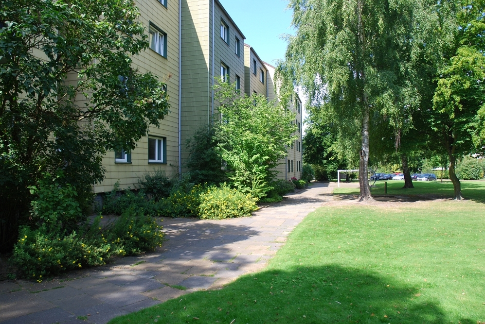 Mehrstöckiges Wohngebäude mit Garten, Bäumen und Gehweg bei Sonnenschein.