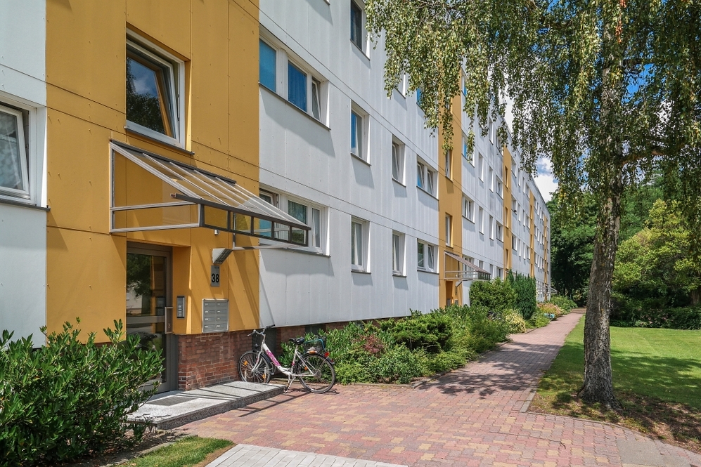 Mehrstöckiges Wohnhaus mit gelb-weißer Fassade und begrüntem Gehweg im Sommer.