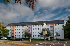 Mehrstöckiges Wohngebäude mit Parkplätzen und Bäumen bei bewölktem Himmel.