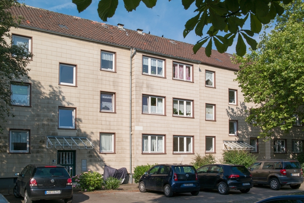 Mehrfamilienhaus mit Parkplätzen und Bäumen, beige Fassade, rote Ziegeldach, ruhige Wohngegend.