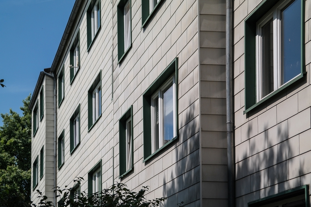 Mehrgeschossiges Wohngebäude mit hell verkleideter Fassade und grünen Fensterrahmen bei Sonnenschein.