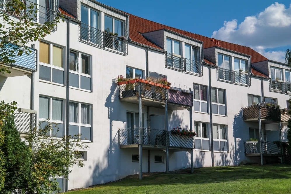 Mehrfamilienhaus mit Balkonen und roten Dachziegeln vor blauem Himmel.