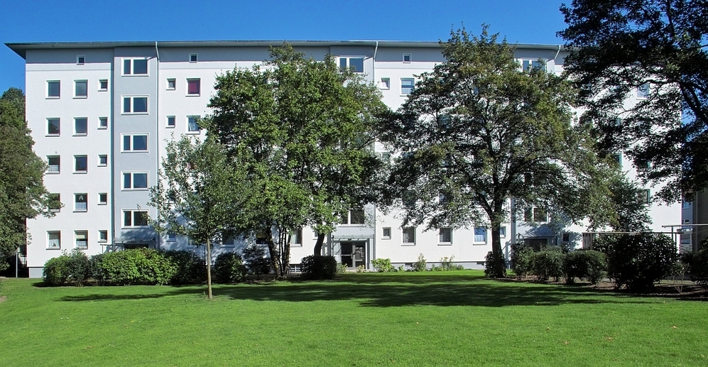 Mehrfamilienhaus mit weißer Fassade, Grünfläche und Bäumen im Vordergrund bei klarem Himmel.