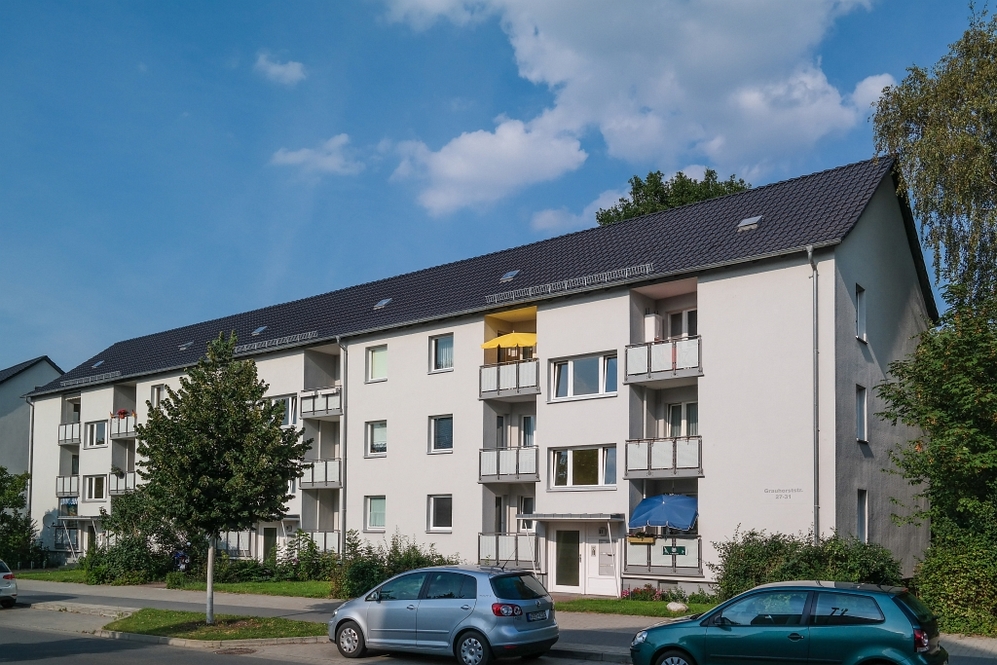 Mehrfamilienwohnhaus mit Balkonen, zwei Parkplätzen und Bäumen an einer Wohnstraße.