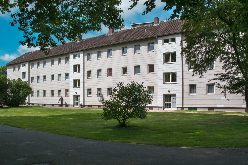 Mehrgeschossiges Wohngebäude mit weißer Fassade, grünem Vorgarten und Bäumen im Umfeld.