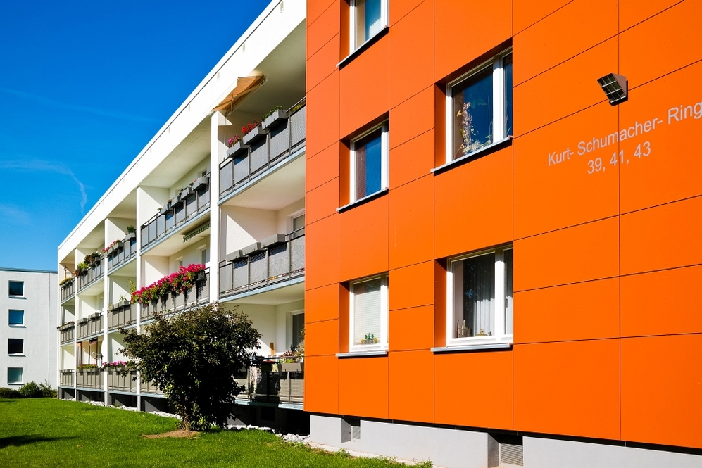 Mehrstöckiges Wohngebäude mit oranger Fassade und Balkonen mit Blumen bei Sonnenschein.