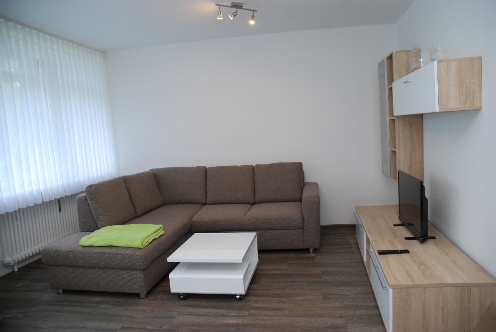 Wohnzimmer mit grauem Ecksofa, weißem Couchtisch und TV auf Holz-Board, helle Vorhänge.