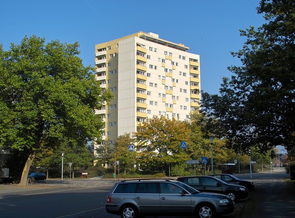 Hochhaus-Wohngebäude mit Balkonen, umgeben von Bäumen und geparkten Autos im Vordergrund.