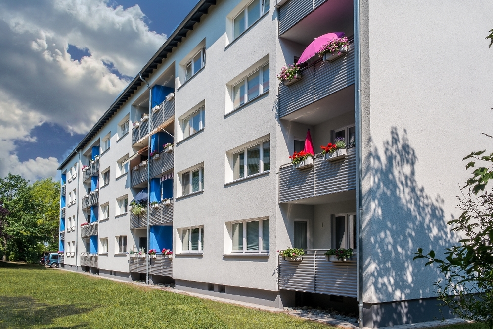 Mehrstöckiges Wohnhaus mit Balkonen und Blumen, umgeben von Grünflächen bei Sonnenschein.