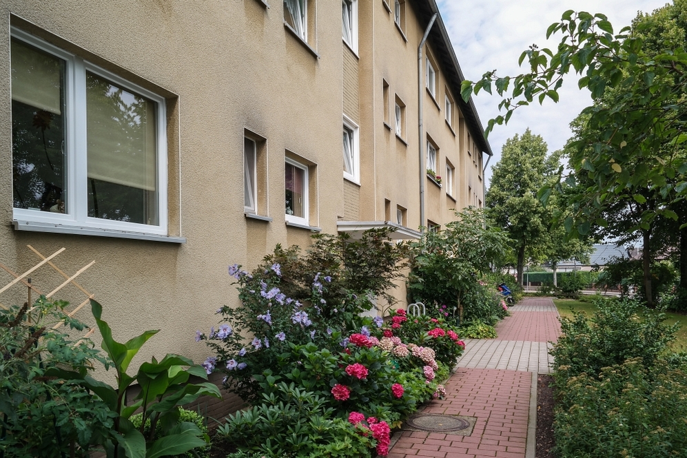 Mehrstöckiges Wohngebäude mit Blumenbeeten und gepflastertem Gehweg im Vorgarten.
