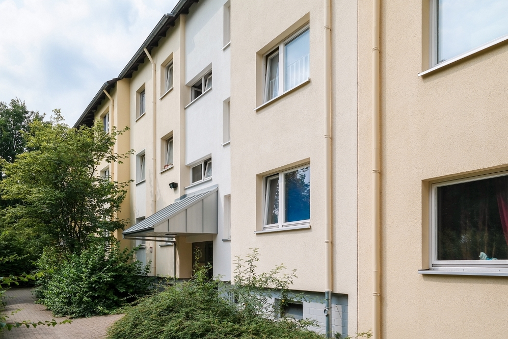 Mehrstöckiges Wohngebäude mit beige-weißer Fassade und begrüntem Eingangsbereich.