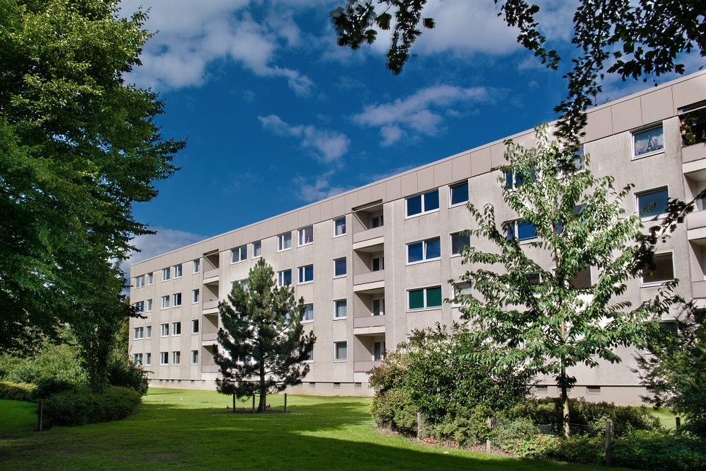 Mehrgeschossiges Wohngebäude mit Grünfläche und Bäumen vor blauem Himmel.