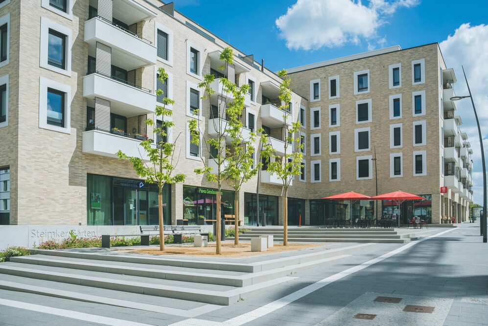Moderne Wohn- und Geschäftshäuser mit Balkonen, Bäumen und Sitzbereichen im urbanen Quartier.