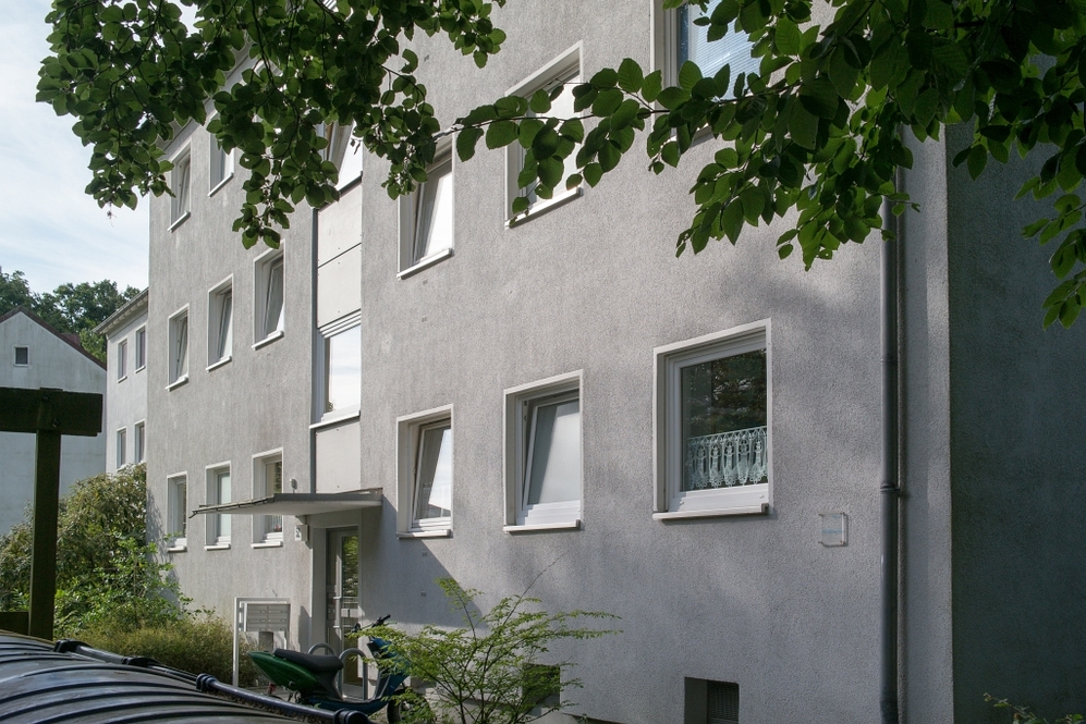 Mehrstöckiges Wohnhaus mit mehreren Fenstern, Eingang und begrüntem Umfeld.
