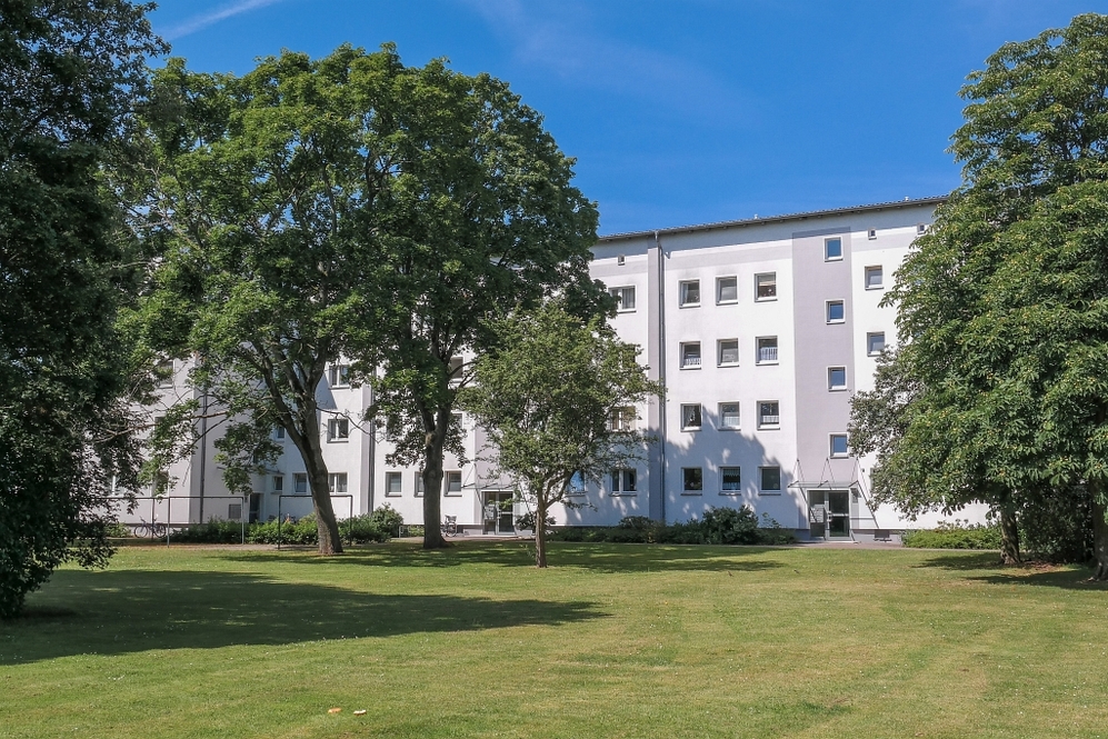 Mehrfamilienhaus mit weißer Fassade, umgeben von großem Rasen und mehreren Bäumen bei klarem Himmel.