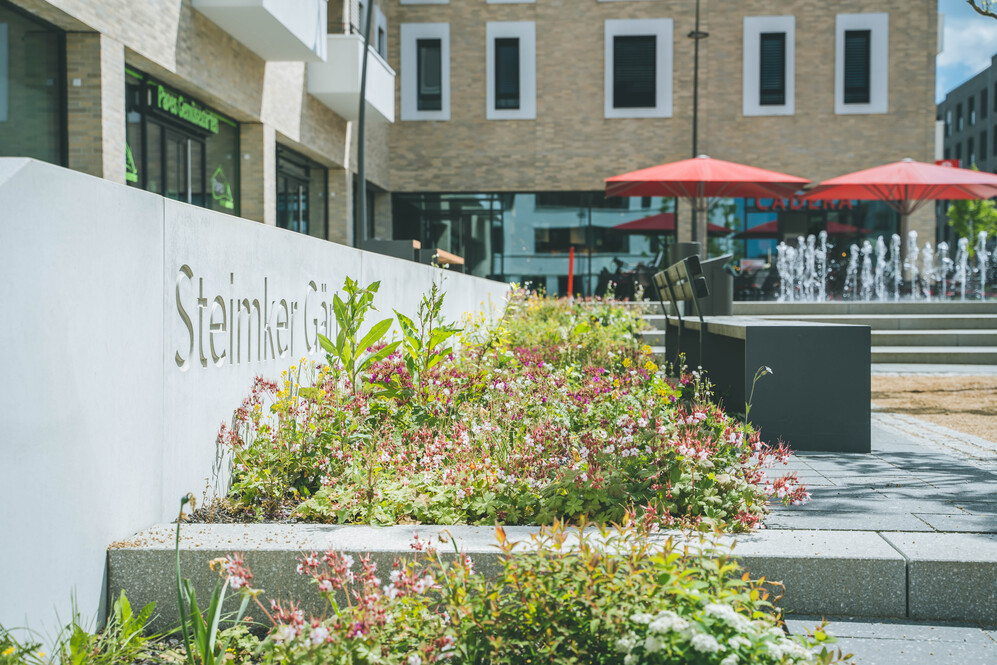 Moderner urbaner Platz mit Sitzbänken, Blumenbeeten und Springbrunnen im Steinmer Garten.