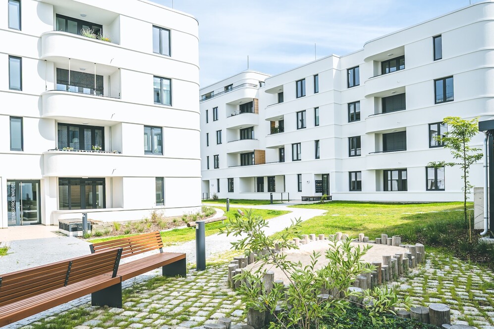 Moderne weiße Wohngebäude mit Balkonen und Grünflächen, Sitzbänken im Innenhof.