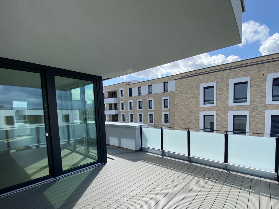 Große Balkonterrasse mit Glasgeländer und Blick auf moderne Mehrfamilienhäuser.
