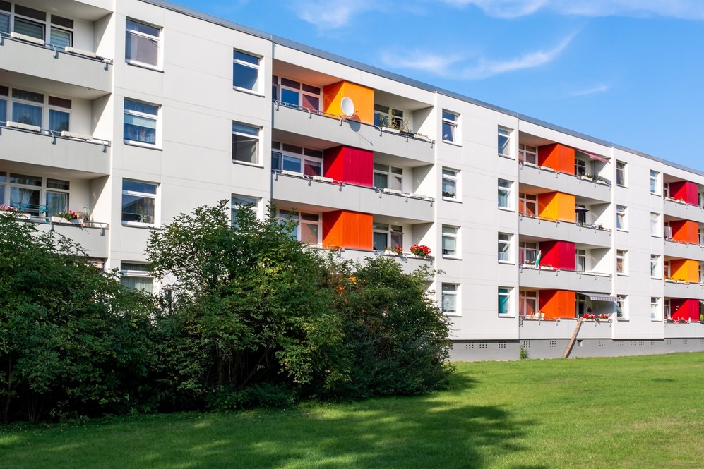 Mehrstöckiges Wohngebäude mit bunten Balkonen und Grünfläche im Vordergrund bei Sonnenschein.
