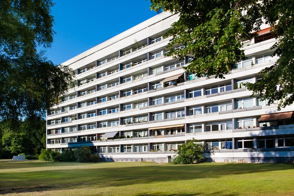 Mehrgeschossiges Wohngebäude mit Balkonen, umgeben von Rasen und Bäumen bei klarem Himmel.