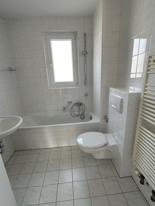 Kleines Badezimmer mit Badewanne, Wand-WC, Waschbecken und Fenster, weiß gefliest.