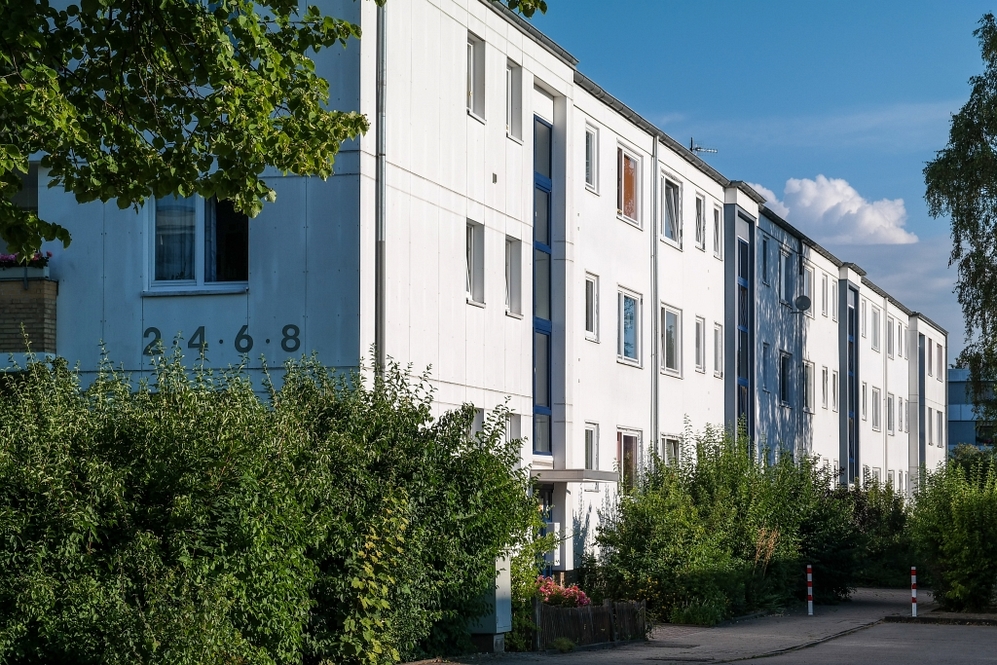 Mehrfamilienhaus mit weißer Fassade, mehreren Fenstern und grüner Bepflanzung im Vordergrund.