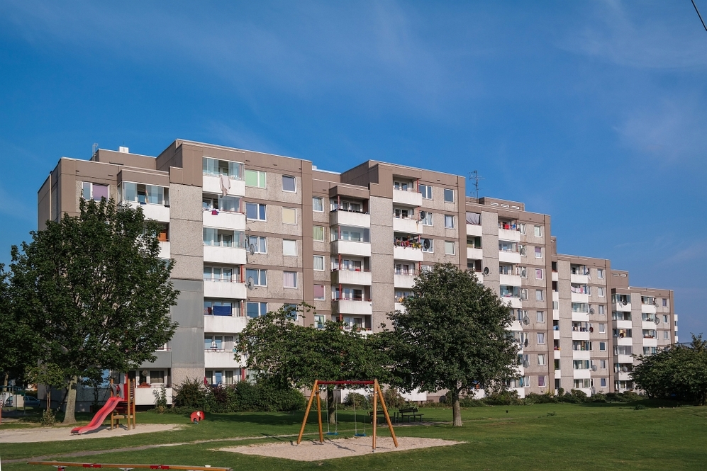 Mehrfamilienhaus mit Balkonen, umgeben von Bäumen und Spielplatz im Vordergrund.