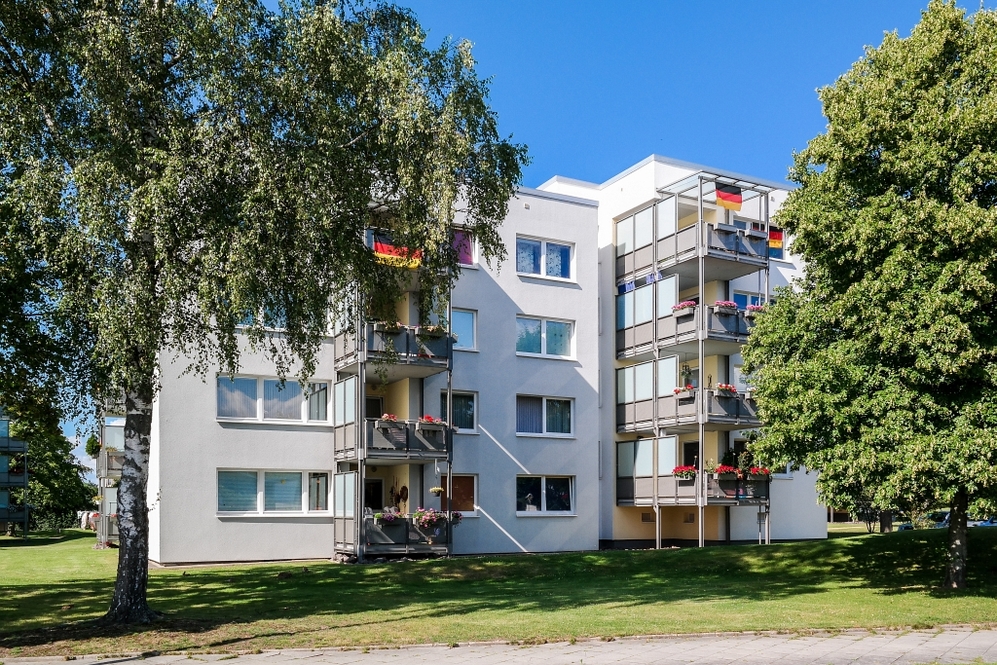 Mehrstöckiges Wohngebäude mit Balkonen, Blumen und deutschen Flaggen, umgeben von Bäumen.
