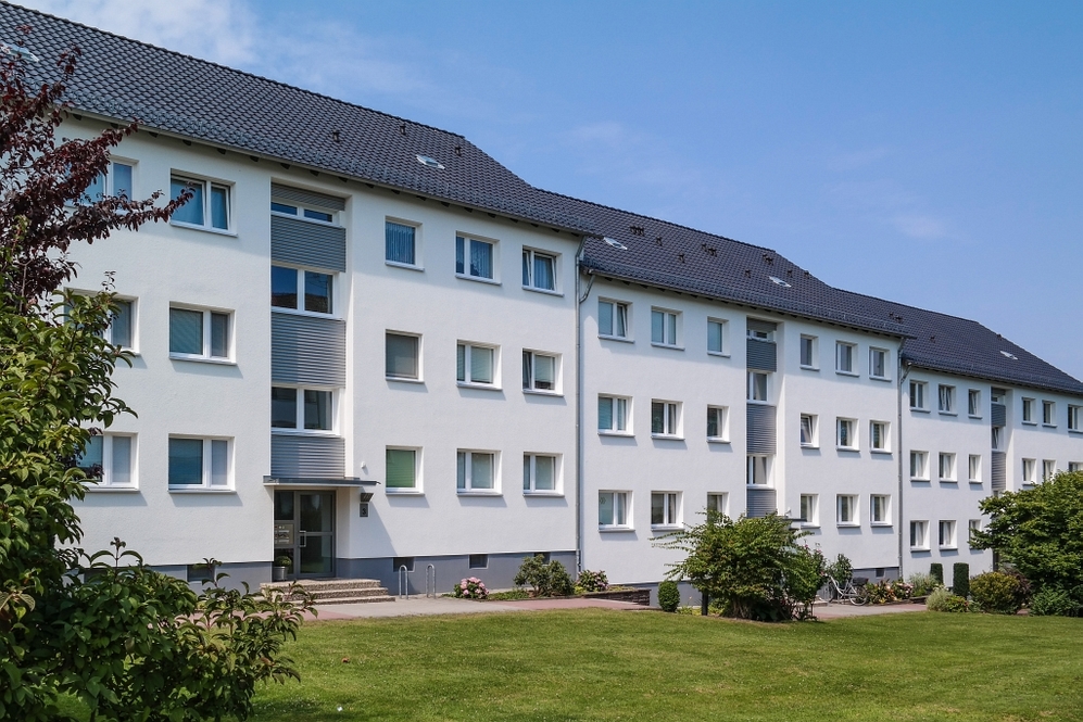 Mehrfamilienhaus mit weißer Fassade, dunklem Dach und gepflegtem Vorgarten bei Sonnenschein.