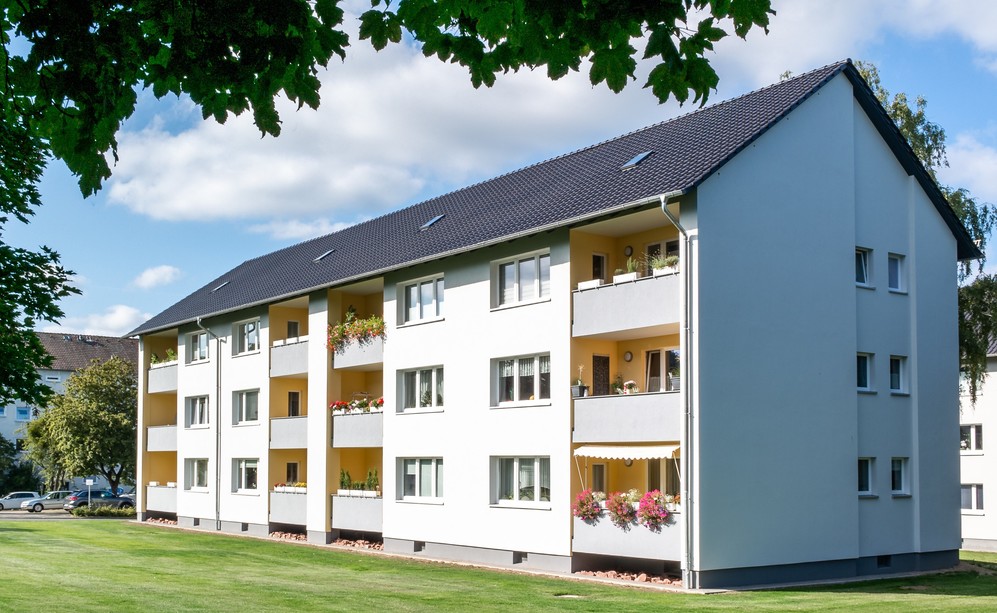 Mehrfamilienhaus mit Balkonen, gepflegtem Rasen und blühenden Pflanzen unter blauem Himmel.